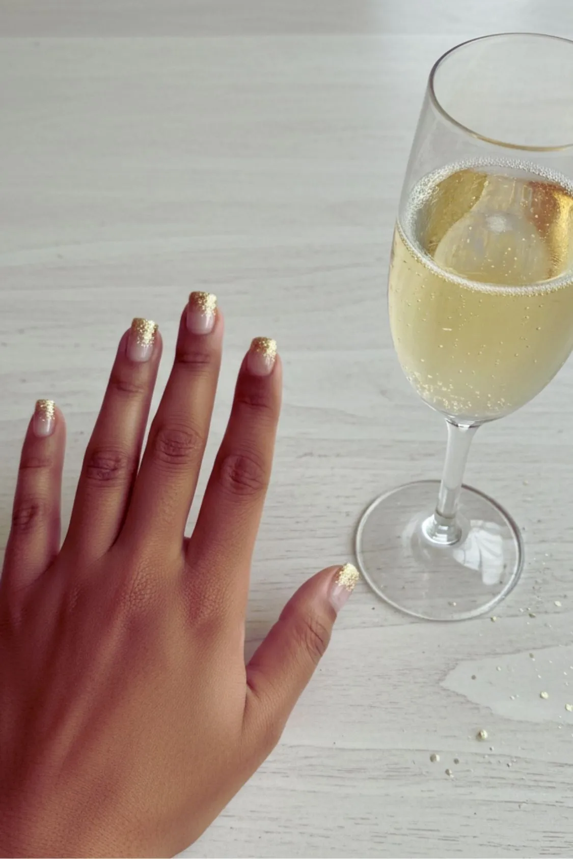 A hand with minimalist champagne glitter nails holding a champagne flute, celebrating the new year.